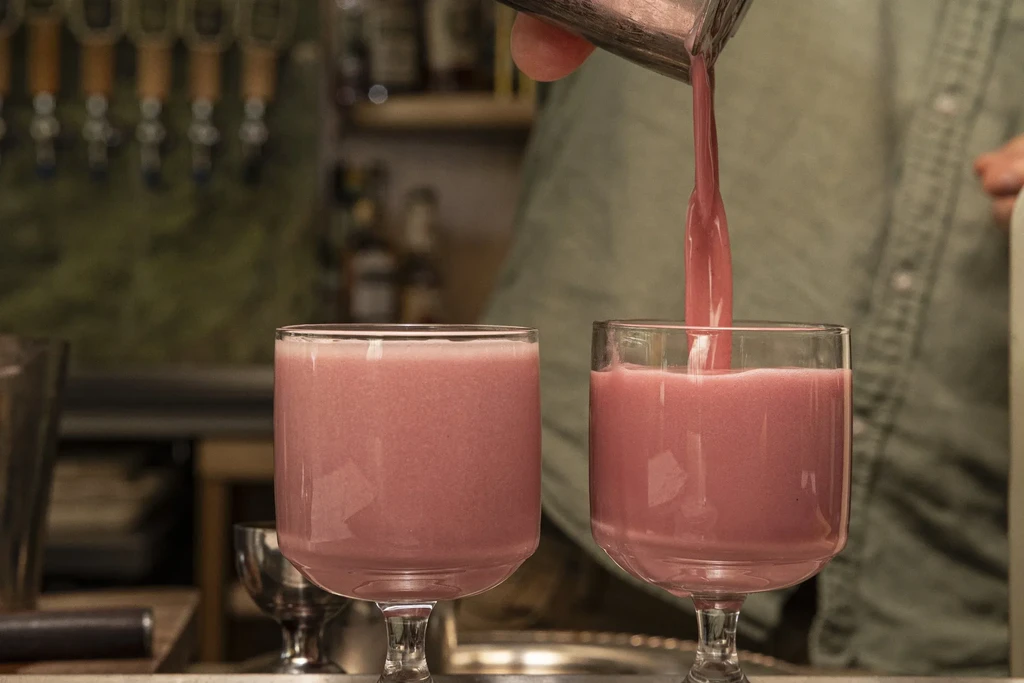 Pink frothy drink being poured into a stemmed glass at a bar.