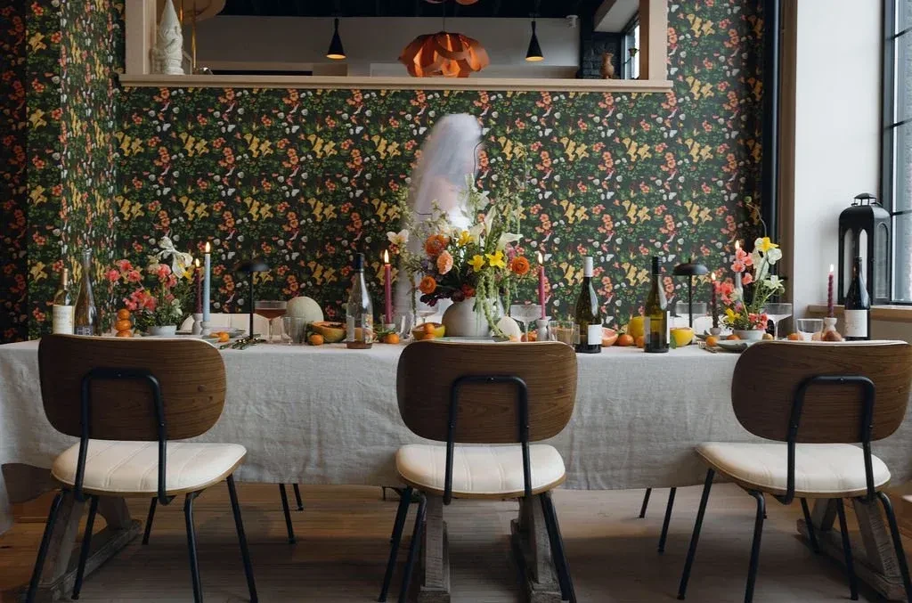 Elaborate dining table with flowers, candles, wine, against floral wallpaper.