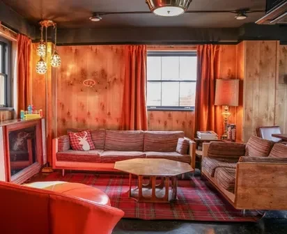 Vintage wood-paneled lounge with orange curtains, striped sofas, and a plaid rug.