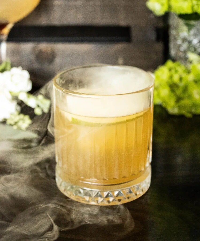 Golden amber cocktail with foam, lime, and smoke in a faceted glass.