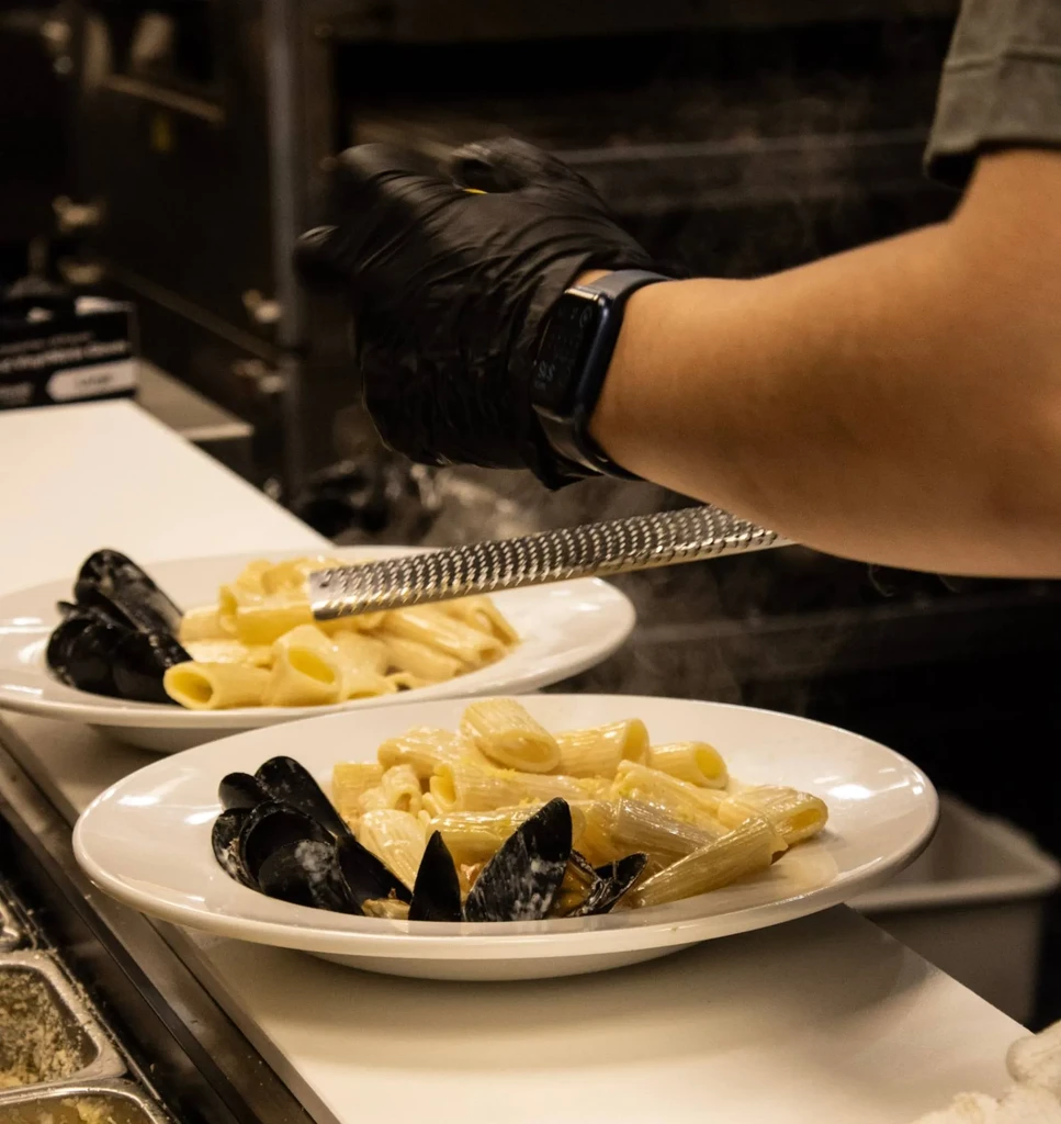 Two plates of hot pasta with mussels, being grated by a gloved hand.