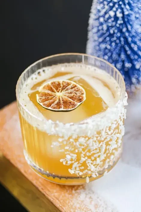 Close-up of golden amber cocktail with coconut rim, lime, and foam.