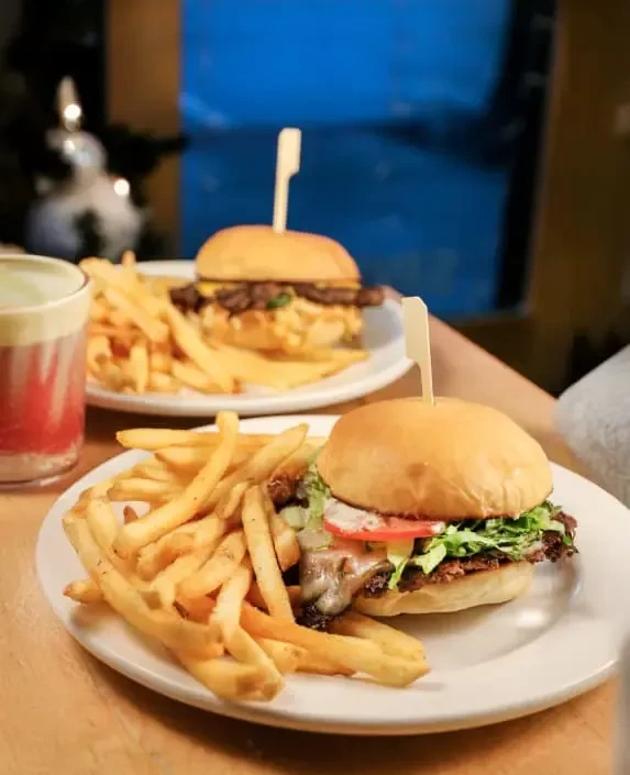 Two plates with cheeseburgers, fries, and a drink on a wooden table.