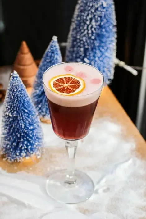 Stemmed glass cocktail, red liquid, pink foam, orange slice, snowy trees.