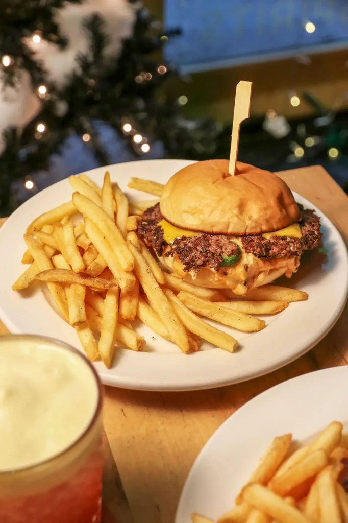Close-up smashburger with melted cheese, fries, and blurred Christmas lights.