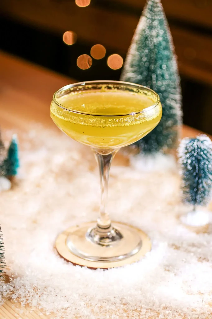 Sugar-rimmed yellow-green cocktail in coupe glass amidst snowy holiday decor.