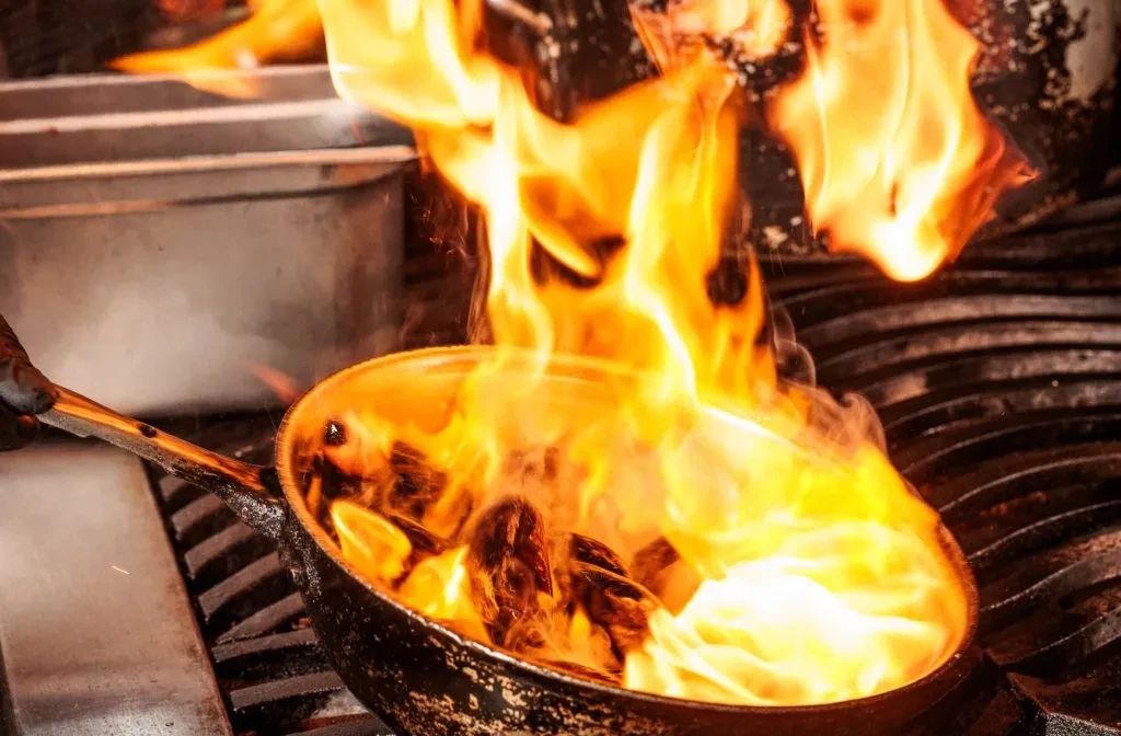 Gloved hand holds black pan with food engulfed in vivid flames.