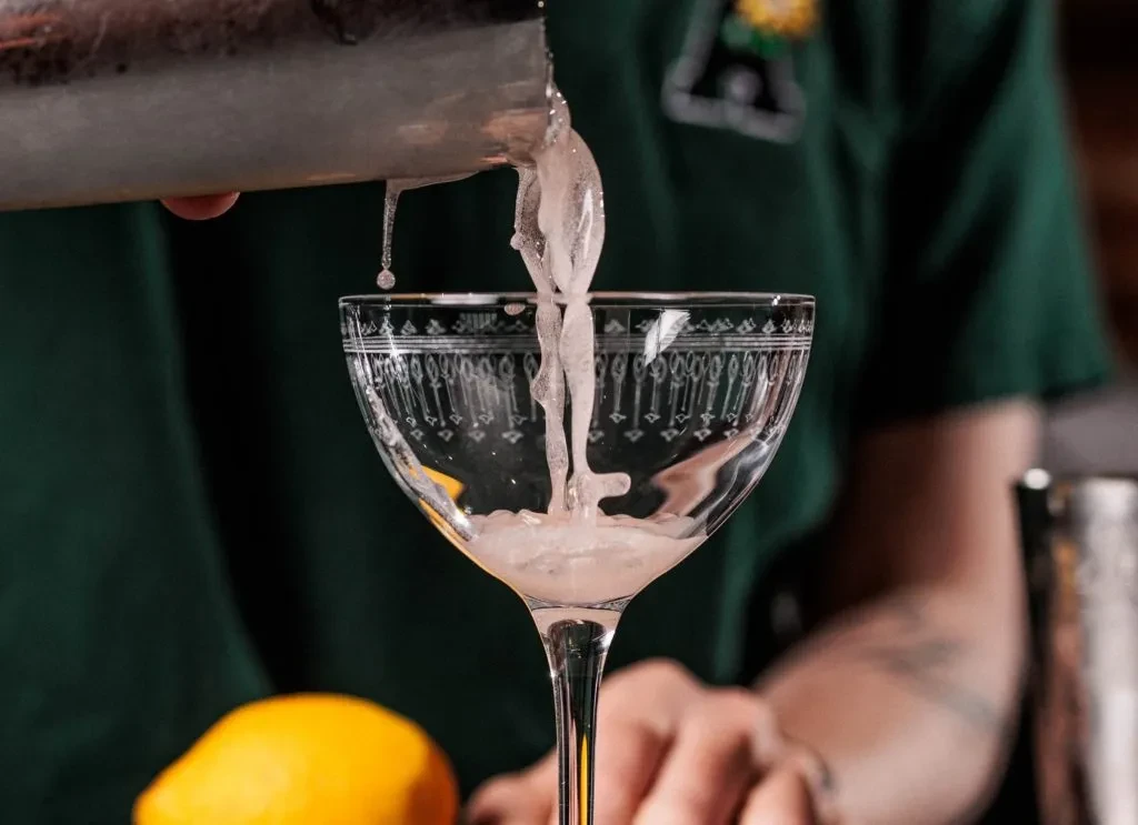 Hand pours frothy cocktail from shaker into etched coupe glass.