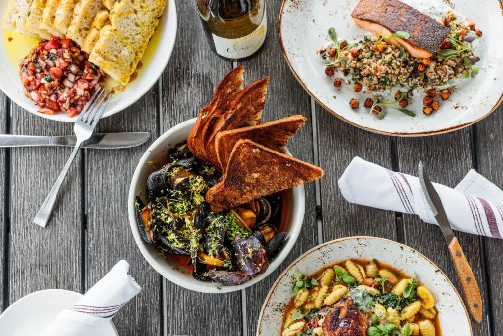 Overhead, a rustic table holds four seafood, pasta dishes, and wine.
