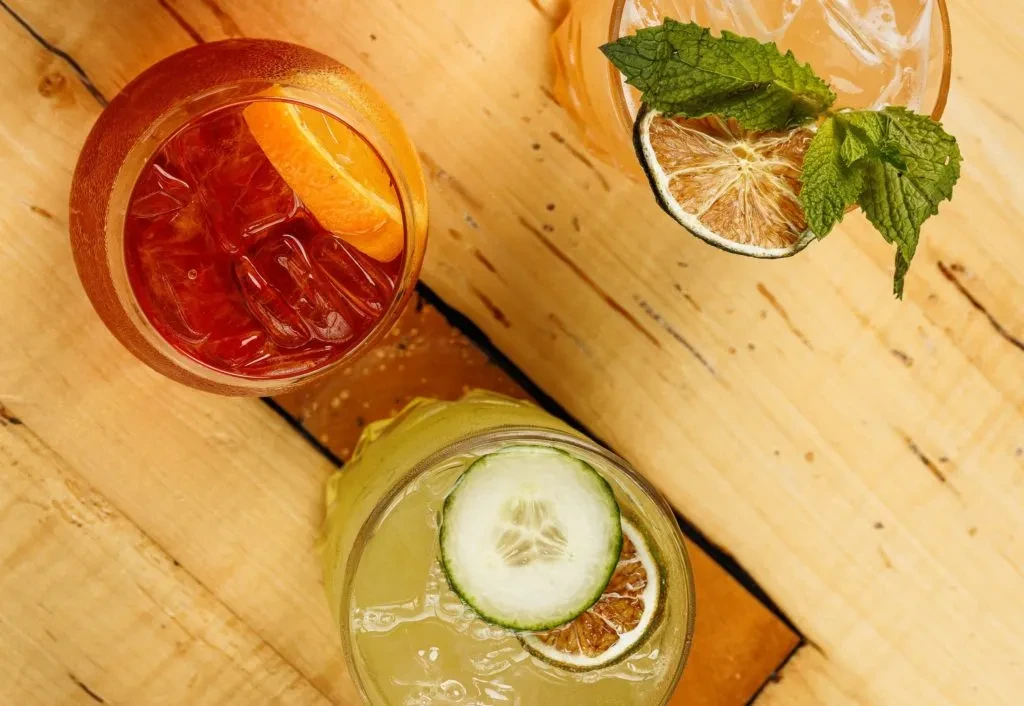 Overhead view of three garnished cocktails on a light wooden table.