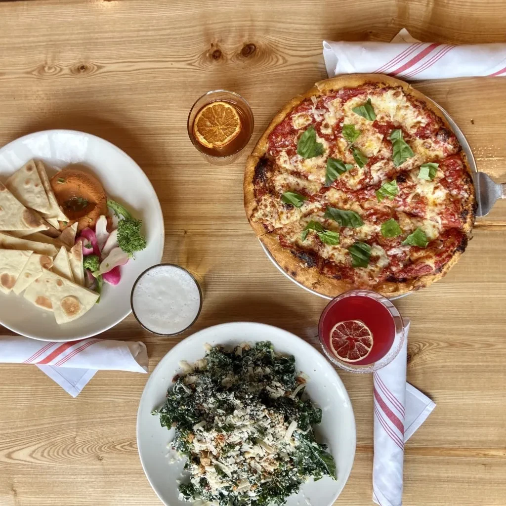Overhead view of pizza, kale salad, hummus, pita, vegetables, and drinks.