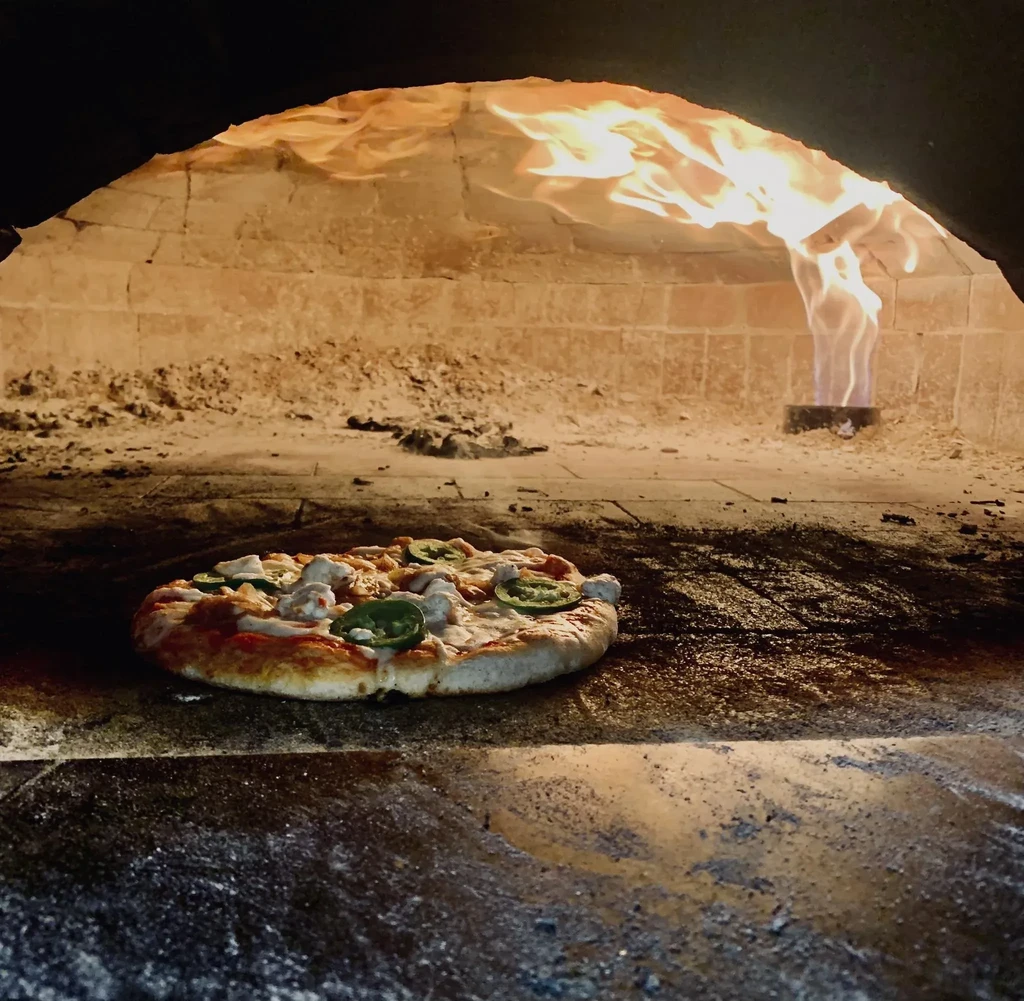 Pizza with cheese and toppings baking in a fiery brick oven.