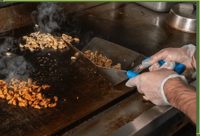 Steam rises as gloved hands cook meat on a flat-top grill.