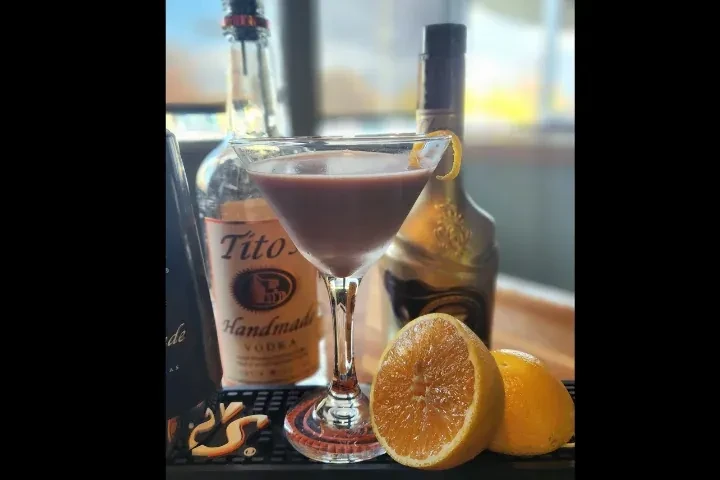 Reddish-brown cocktail in martini glass with lemon twist, oranges, and vodka.