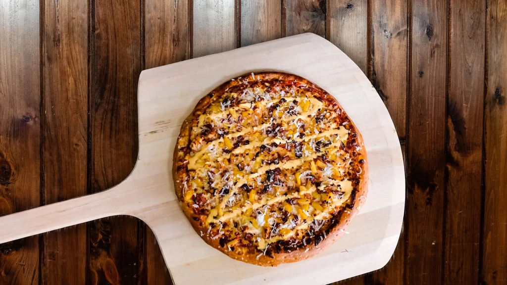 Pineapple pizza with cheese and sauce drizzle on a wooden peel.