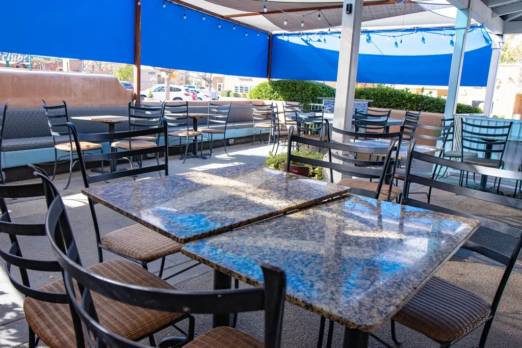 Empty, sunny dining patio with tables, chairs, blue awnings, and lights.