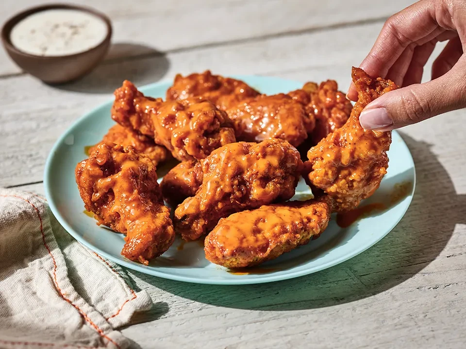 Hand picks one of nine buffalo wings from blue plate on wood.