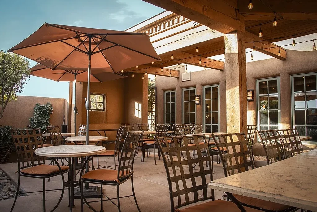 Empty outdoor patio with adobe walls, tables, string lights, and umbrella.