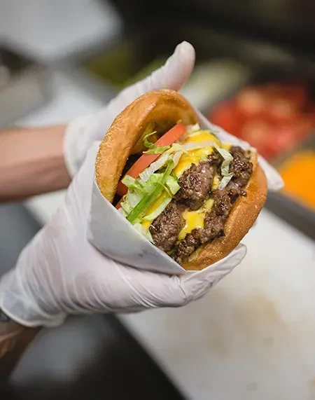 Gloved hand holds a double cheeseburger with cheese, lettuce, and tomato.