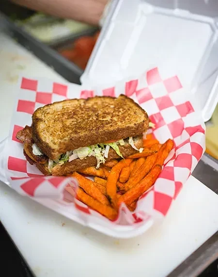 Grilled sandwich halves, sweet potato fries in a checkered takeout container.