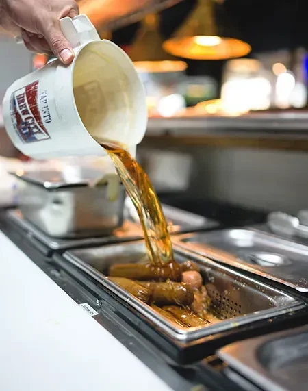Hand pours liquid from "BREW CO TAP 1970" mug over hot dogs.