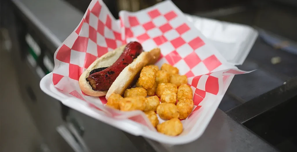 Grilled hot dog in bun with golden tater tots in a container.