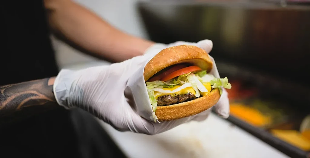 Gloved hands holding a fresh cheeseburger wrapped in paper.