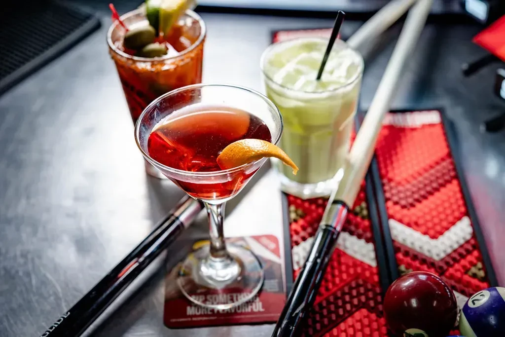 Three cocktails on a bar next to pool cues and balls.
