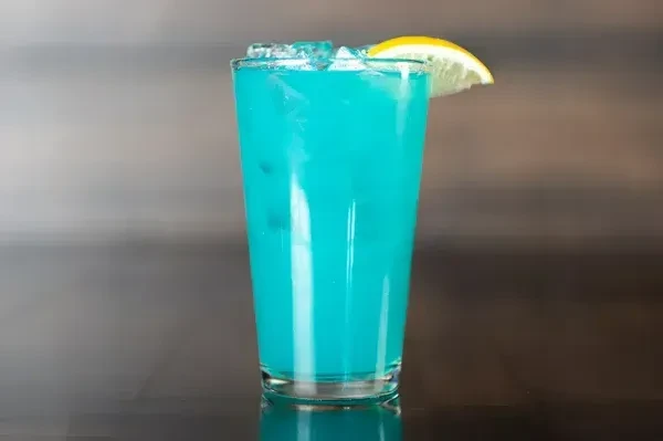 Tall glass of blue drink with ice and a lemon wedge.