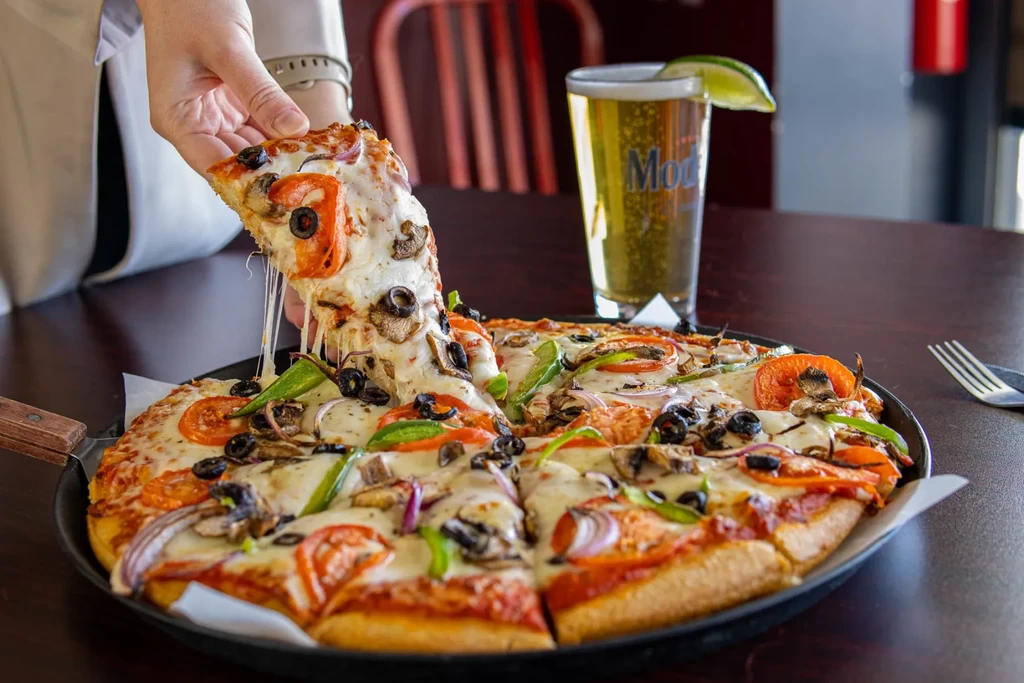Close-up of pizza, hand lifting slice with cheese pull, beer and cutlery.