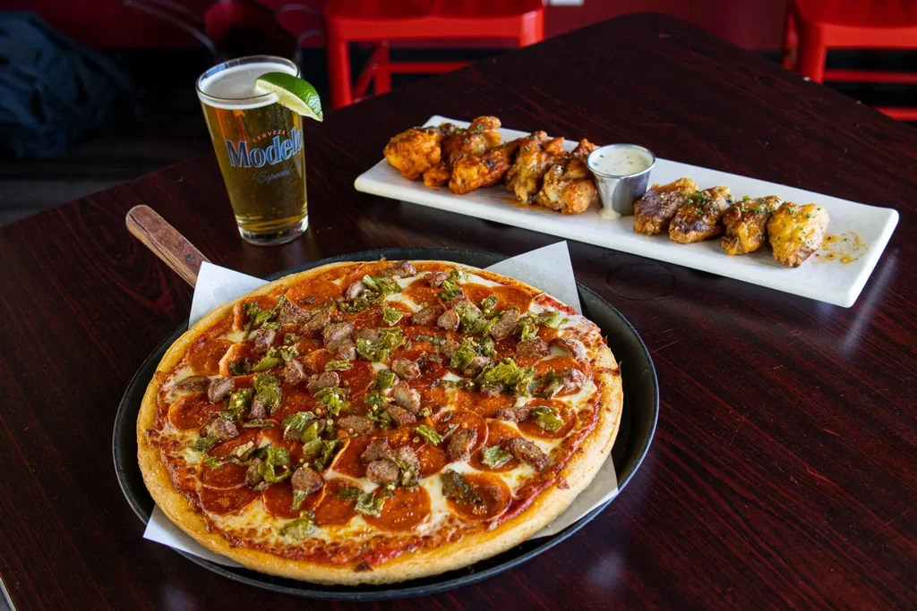 Skillet pizza, chicken wings, Modelo beer, and sauce on a dark table.