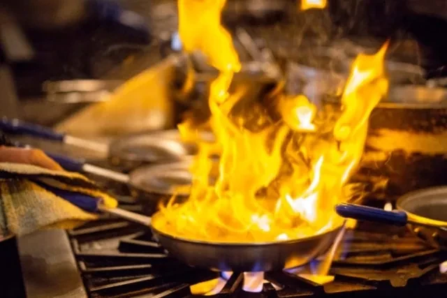 A chef's hand holds a pan flambe cooking with bright flames.
