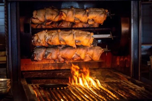 Golden brown rotisserie chickens roasting over open flames in an industrial oven.