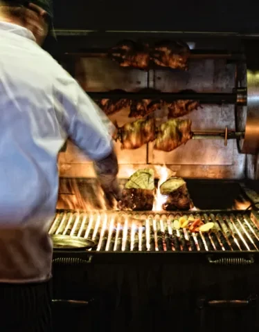 Chef roasts chickens, grills meats and vegetables on open fire.