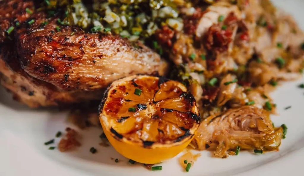 Plated roasted meat, green herb garnish, charred lemon, and side vegetables.