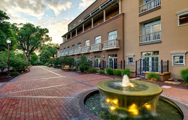 Evening view of an illuminated fountain, building, and tree-lined brick path.