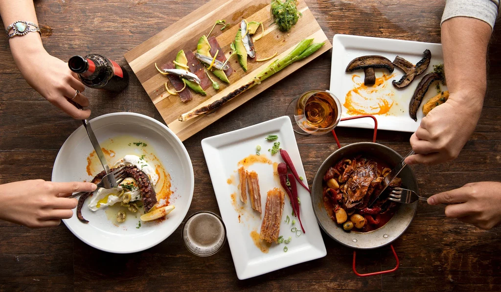Hands cutting and eating gourmet dishes and drinks on a rustic table.
