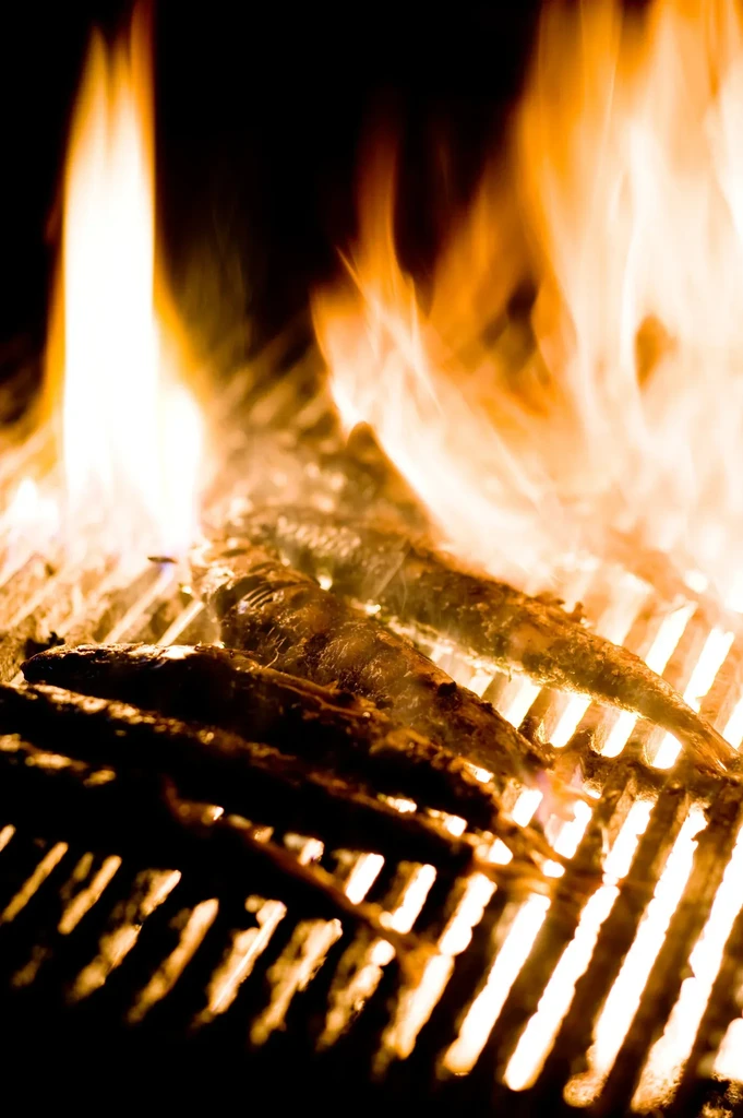 Small, dark fish grilling over vibrant orange and yellow flames.