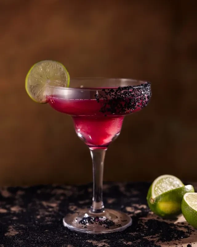 Magenta margarita cocktail with black sugar rim and lime.