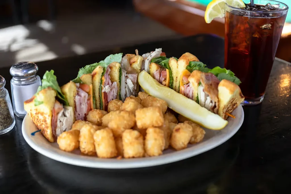 Well-lit club sandwich meal with tater tots, pickle, and iced drink.