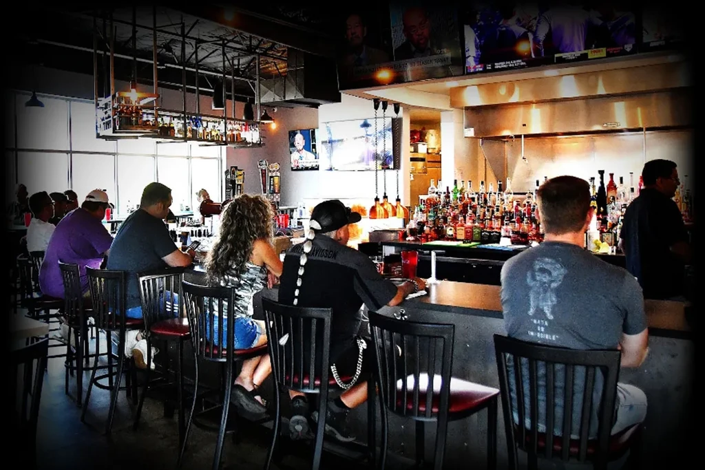 Busy bar interior with a well-stocked counter, patrons, and TVs.