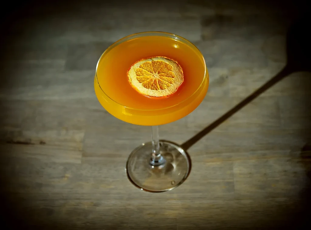 Overhead orange-yellow cocktail with dried orange in stemmed glass on wood.