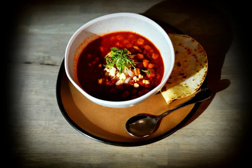 Overhead close-up of red stew with herbs, tortilla, and spoon.