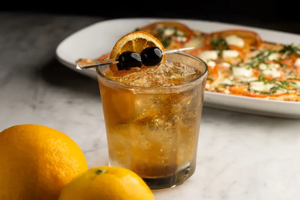 Amber cocktail with dried orange and cherries, blurred Caprese and oranges.