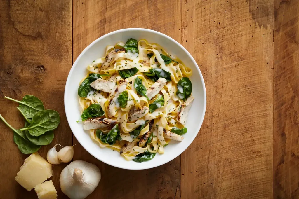 White bowl of chicken Alfredo pasta, raw ingredients on wooden surface.