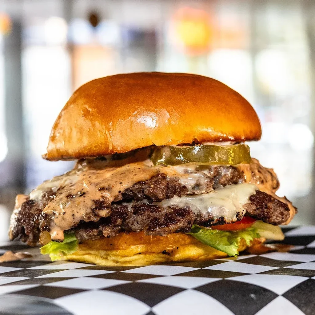 Stacked burger with brioche bun, beef patties, melted cheese, pickle.