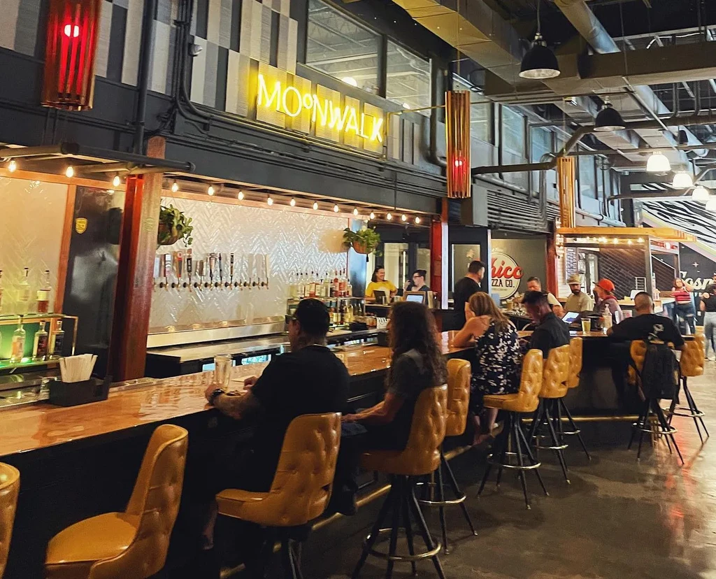Lively bar interior with patrons, yellow stools, and "MOONWALK" neon sign.