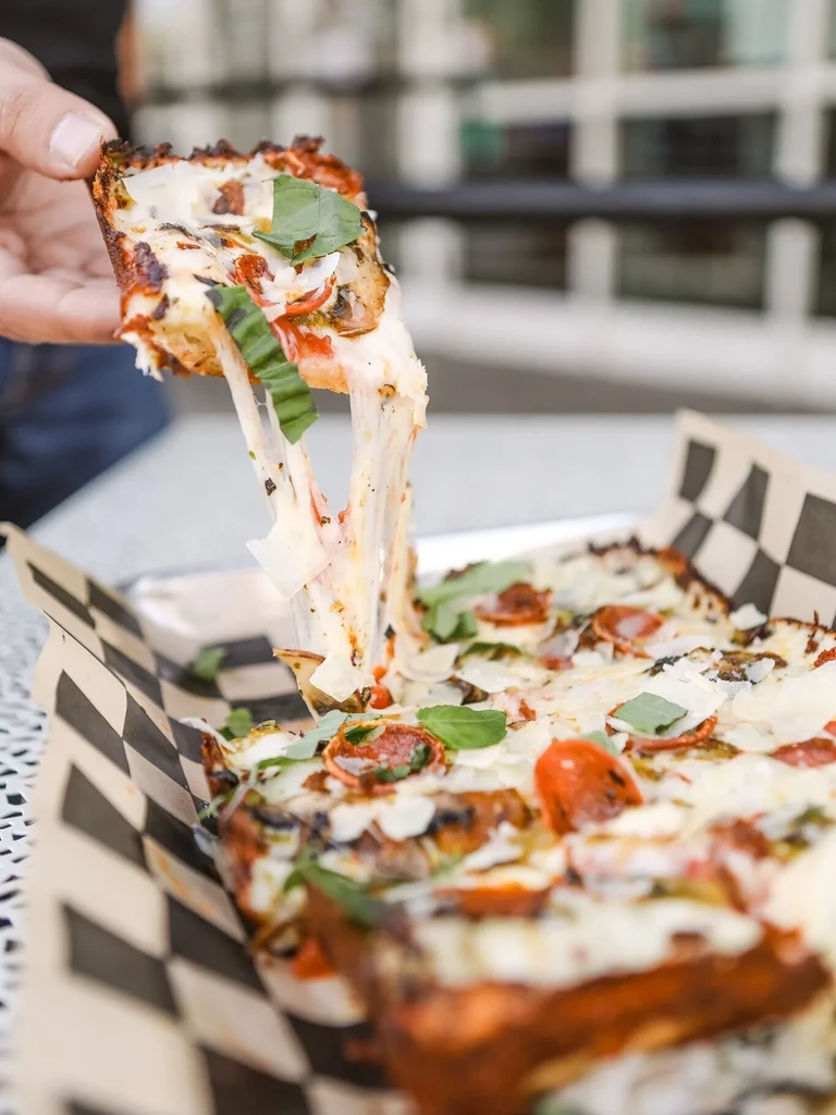 Hand lifts deep-dish pizza slice showing cheese pull, pepperoni, and basil.