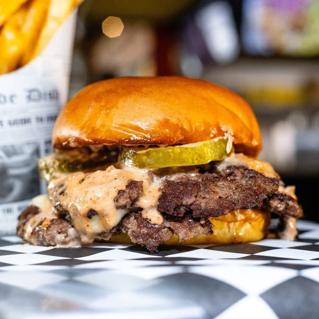 Double cheeseburger with sauce, pickles, and fries on checkered paper.