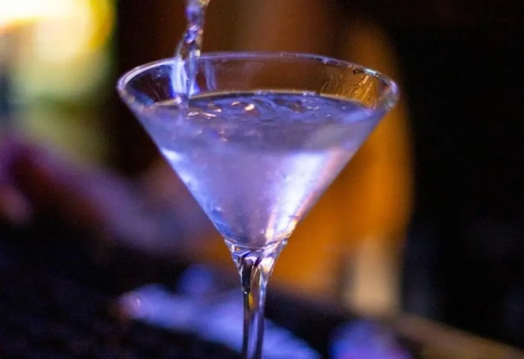 Bluish-purple cocktail pouring into a martini glass in a dim bar.
